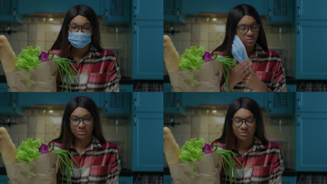 戴着眼镜的年轻非洲裔美国妇女脱下面部防护面罩，看着坐在厨房桌子上的相机，带着新鲜食物的纸袋。特写