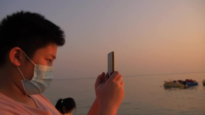 快乐的亚洲男孩的剪影通过智能手机在海滩上拍摄照片和自拍，度假期间日出，生活方式概念。
