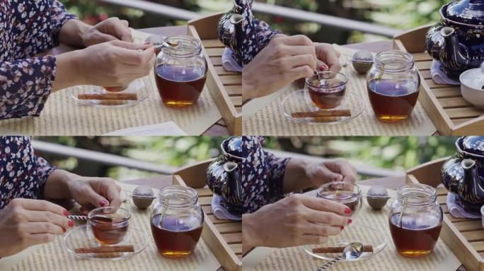 在户外露台的桌子上，年轻女子的手将蜂蜜天然甜味剂倒入热茶杯中的特写镜头