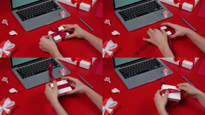 女人的手打开礼品盒，解开蝴蝶结。带笔记本电脑、礼物、心形和情人节的红色桌子的俯视图。浪漫的心情。情人