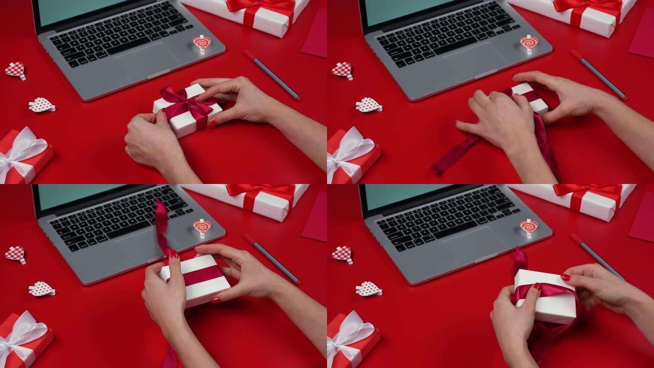 女人的手打开礼品盒，解开蝴蝶结。带笔记本电脑、礼物、心形和情人节的红色桌子的俯视图。浪漫的心情。情人