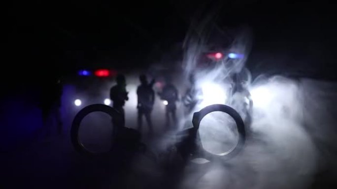 警察在晚上突袭，你被逮捕了。手铐的轮廓，背面有警车。在有雾的背景下闪烁的红色和蓝色警灯的图像。选择性