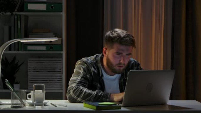 晚上，疲惫的男人坐在笔记本电脑附近的家庭办公室工作室的办公桌前睡觉。远程工作人员醒来，喝咖啡，然后继