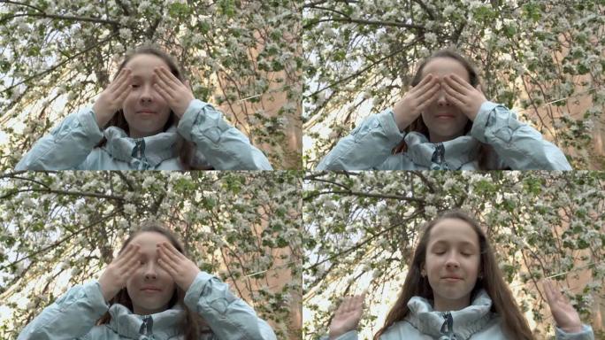 可爱的十几岁的女孩在春天的日子里站在后院的一棵盛开的树的背景下玩耍。她闭上眼睛，用手睁开眼睛，看着镜