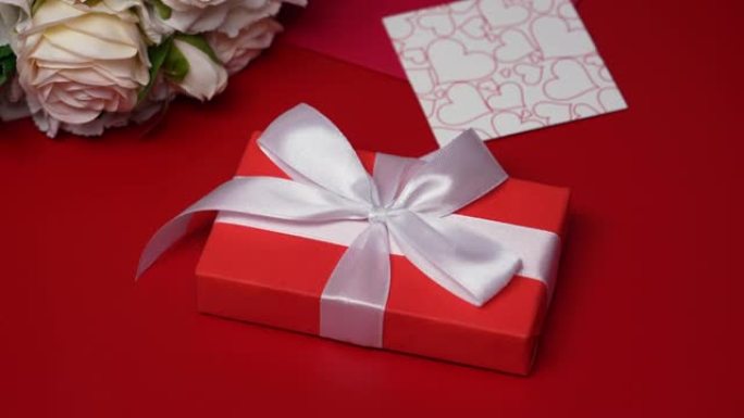 带有白色丝带蝴蝶结的礼品盒落在红色桌子上。节日背景，带有情人节、一束玫瑰、一个信封、情人节和装饰性的