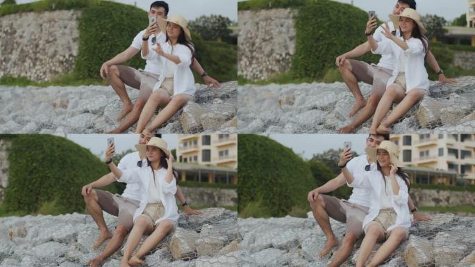 快乐的亚洲夫妇使用智能手机在美丽的日落时坐在海边的石头上拍照和自拍。浪漫情侣在度假周末旅行。