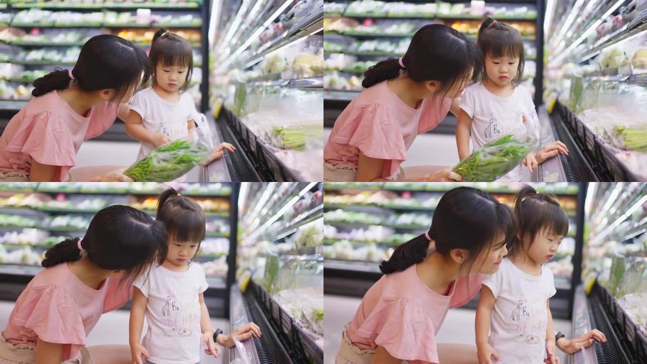 年轻的亚洲美丽的母亲拿着菜篮子，带着她的孩子在超市散步。她正在选择从架子上捡起来的绿色沙拉蔬菜，然后