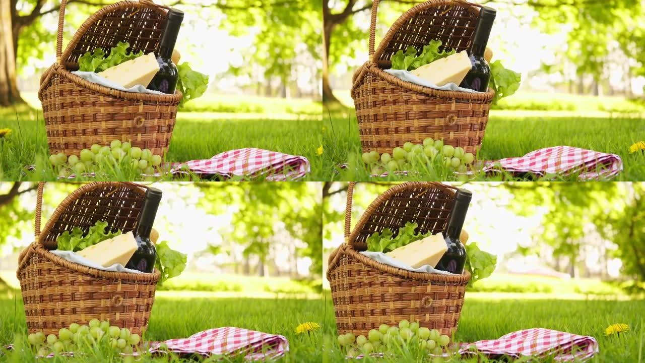 夏季公园外面绿草丛上的红方格桌布上的柳条野餐篮配奶酪和葡萄酒