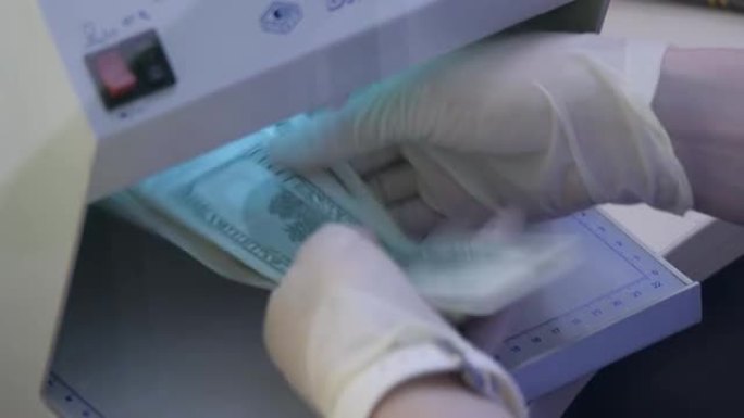 银行员工或货币兑换业务员在货币检测器上的紫外线下检查一百美元钞票中的现金。戴上防护手套，在紫外线探测