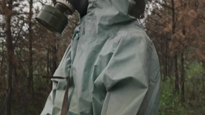 一名男子穿着二战危险品套装和防毒面具，手里拿着一个柜台，在森林里扫描有害物质。原始视频记录