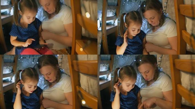 母亲在坐在客厅的帐篷里玩游戏和拍照时，教女儿使用现代智能手机。