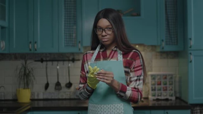 千禧一代非洲裔美国妇女厌倦了家政服务，脱下黄色橡胶手套站在蓝色厨房里。在家穿围裙的女人。