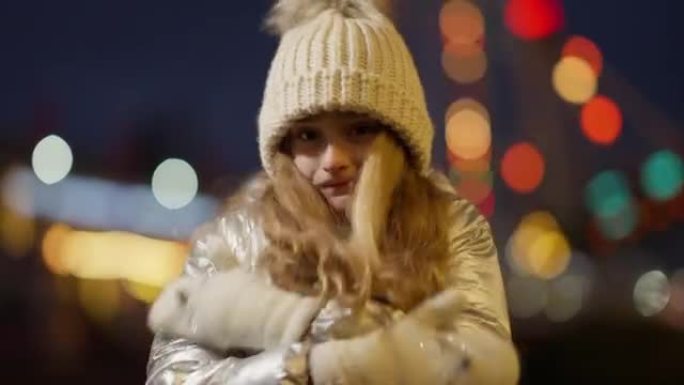 在装饰的夜城背景下，被冻结的高加索女孩在慢动作中摩擦手在户外变暖，新年灯。快乐可爱的孩子在户外庆祝圣