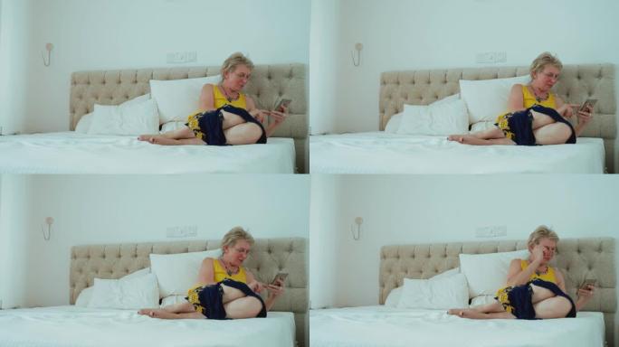 55岁的成熟女性坐在明亮整洁的现代酒店房间的床上时，会用智能手机发短信。