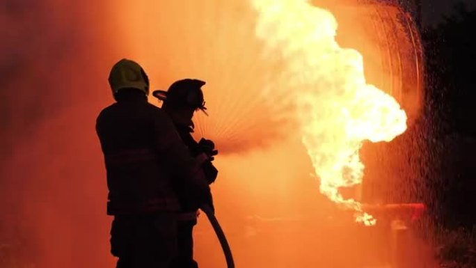 后视图消防队员在完整的装备操作消防水带在消防站在晚上。