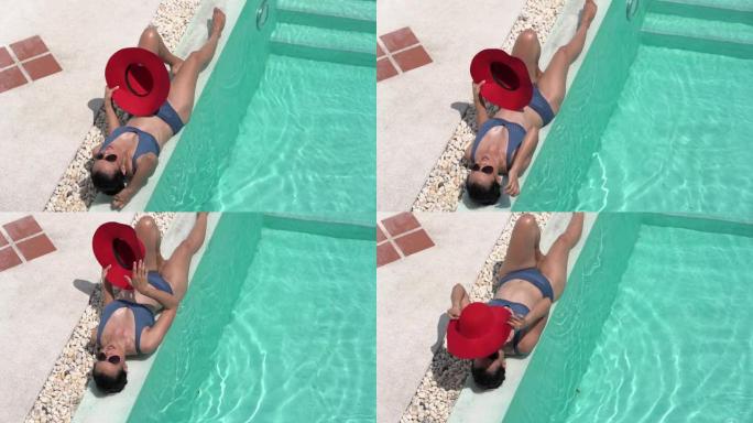 年轻的美女躺在水疗度假酒店的游泳池旁放松。炎热的夏日和明亮的阳光。享受晒黑和放松与大红帽子盖面部保护