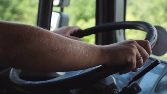 在夏日驾驶卡车时，卡车司机的男性手握着一个大方向盘。卡车司机骑到乡间道的目的地。物流和运输的概念。关