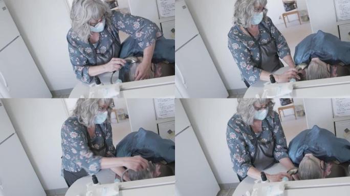 一名专业护理人员造型师的手持照片在家里洗头时，在厨房的水槽里洗了一名白人老年妇女的白发和白发