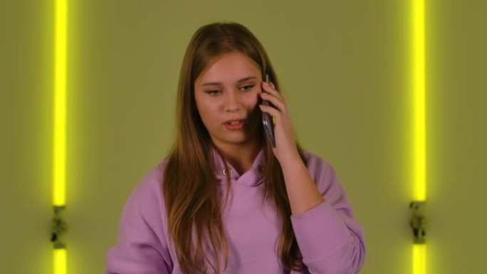 年轻可爱的女人的肖像正在为手机聊天，变得不高兴。穿着紫色连帽衫的长发时装模特在明亮的霓虹灯背景下摆姿