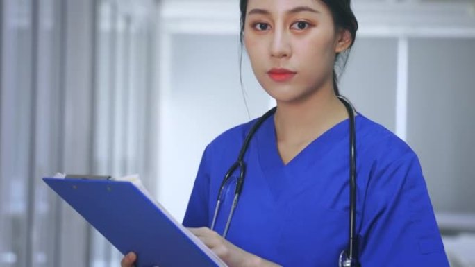 年轻漂亮的亚洲女医生的肖像，穿着蓝色制服，配有听诊器，站在医院走廊上拿着剪贴板检查报告，一边看相机