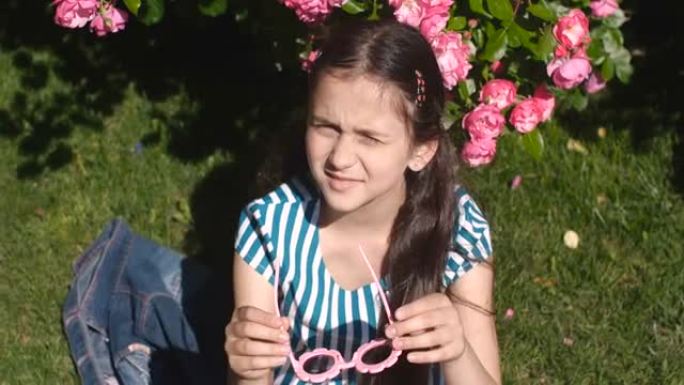 八岁的白人女孩，穿着条纹t恤的长发，坐在草地上，在刺眼的阳光下眯着眼睛，在开花的玫瑰丛中戴上眼镜。4