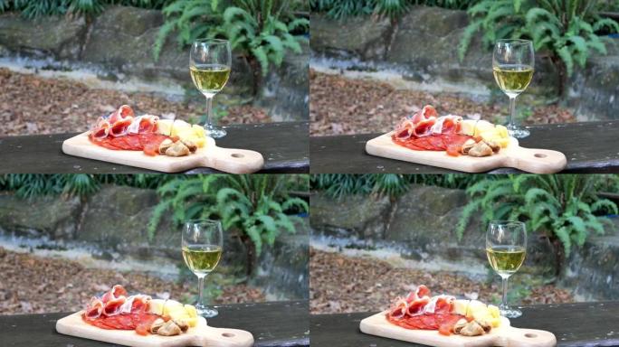 各种奶酪和熟食拼盘的镜头，包括Gouda和Camembert，混合干果和一杯热带花园背景的白葡萄酒。