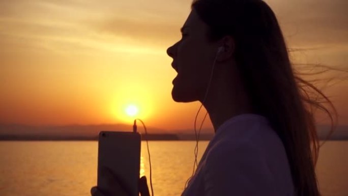 女人的剪影通过手机上的应用程序在慢动作的湖上日落时用耳机听音乐