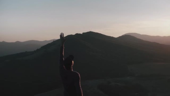 一名年轻人站在夕阳的光芒中，举起手来对一架无人机进行航测，该无人机在他周围绕着一圈飞行，靠山全景