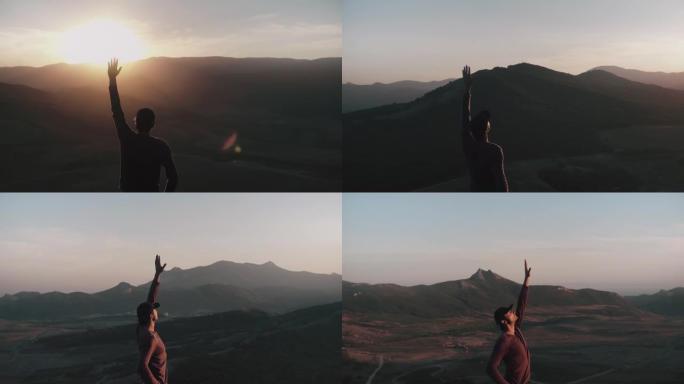 一名年轻人站在夕阳的光芒中，举起手来对一架无人机进行航测，该无人机在他周围绕着一圈飞行，靠山全景