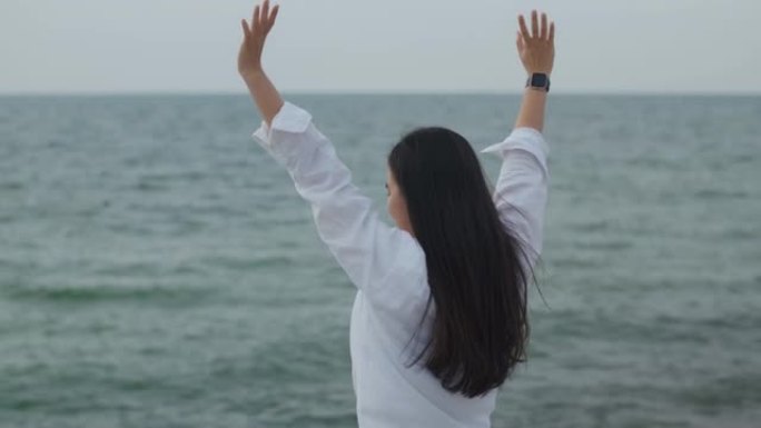 快乐的亚洲女孩庆祝节日周末举起双臂成功。白人女性旅行者站在海滩上，看着海景。