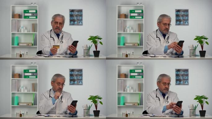 穿着白色医用外套的老年男性医生使用手机和信用卡进行在线购买。白发和眼镜的医生坐在医院办公室的桌子旁。