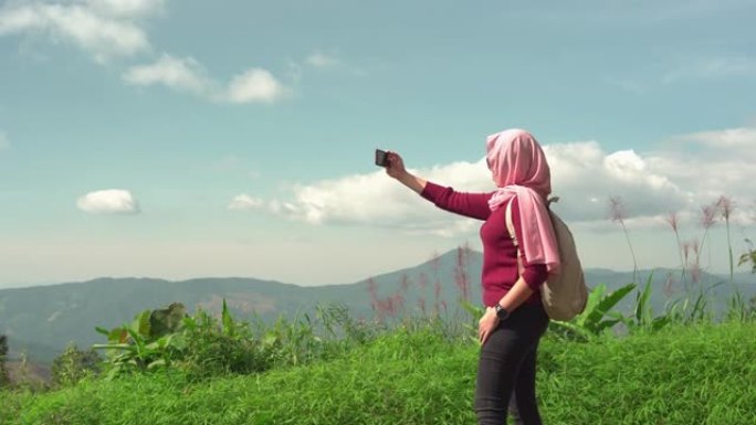 东南亚穆斯林妇女，穿着冬衣，头巾，背包客在智能手机上拍摄全景，看着森林，感受幸福，放松，享受。旅行、