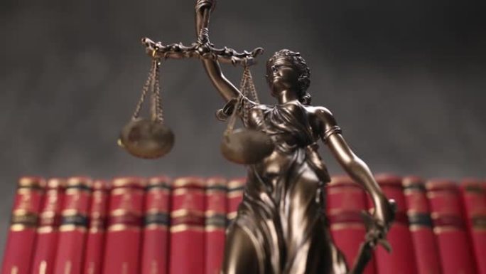 以蒙眼女神为代表的国际法律青铜象征，在一排红书前缓缓旋转，手中握着象征法律两大渊源公正与权力的平衡