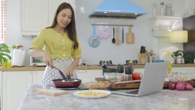 一个美丽的亚洲女孩通过看着如何用快乐的脸用笔记本电脑做早餐来做早餐。在她自己的房子里