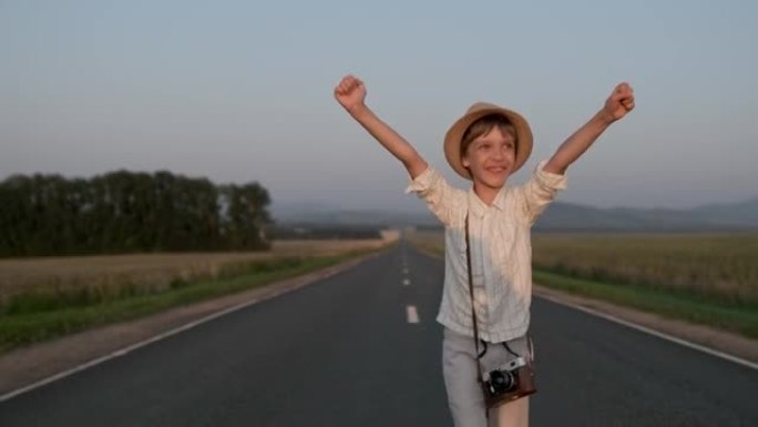 带着相机跳跃的小微笑快乐男孩在夏天的乡村道路上庆祝旅行假期