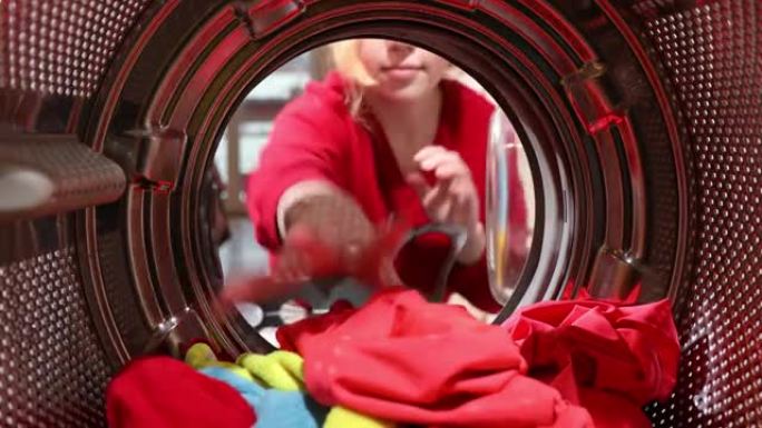 从洗衣机滚筒内看，一名年轻女子从baske装载脏衣服。从洗衣机里拿出衣服。