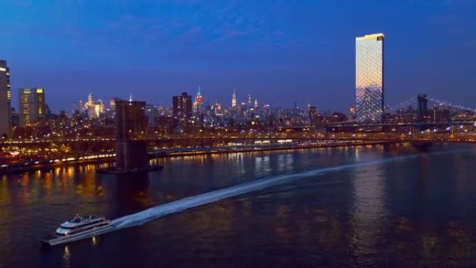 水上渡轮在曼哈顿中城前经过，在夜间照明。带有平移摄像机运动的无人机视频。航拍无人机视频具有向后和向下