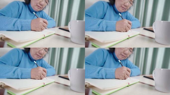 亚洲高级女性在个人日记上写回忆故事，老年人视力问题，戴眼镜，用铅笔在纸质笔记本上写下，呆在家里，家庭