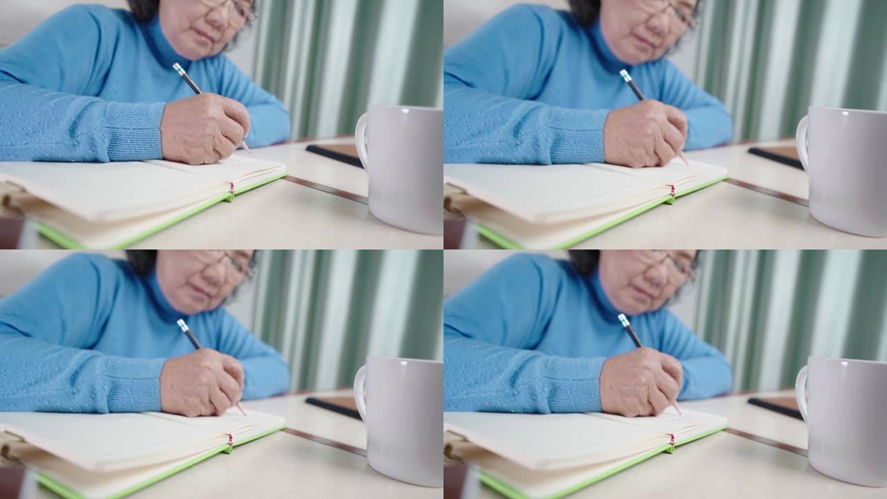 亚洲高级女性在个人日记上写回忆故事，老年人视力问题，戴眼镜，用铅笔在纸质笔记本上写下，呆在家里，家庭