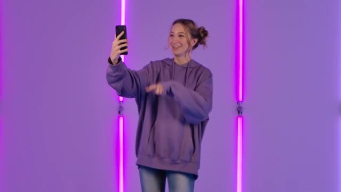 年轻迷人的女人的肖像使用手机聊天视频聊天并欢欣鼓舞。穿着紫色连帽衫的时装模特在黑暗工作室明亮的霓虹灯