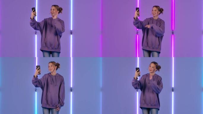年轻迷人的女人的肖像使用手机聊天视频聊天并欢欣鼓舞。穿着紫色连帽衫的时装模特在黑暗工作室明亮的霓虹灯