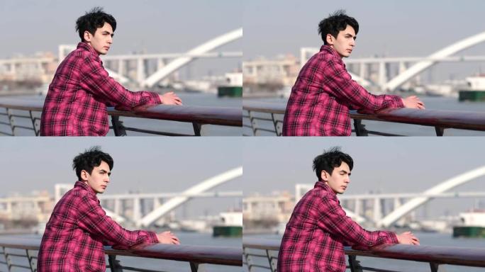 坐在黄浦河河边的英俊中国年轻人的侧视图，带有桥梁和城市背景，4k慢动作镜头。
