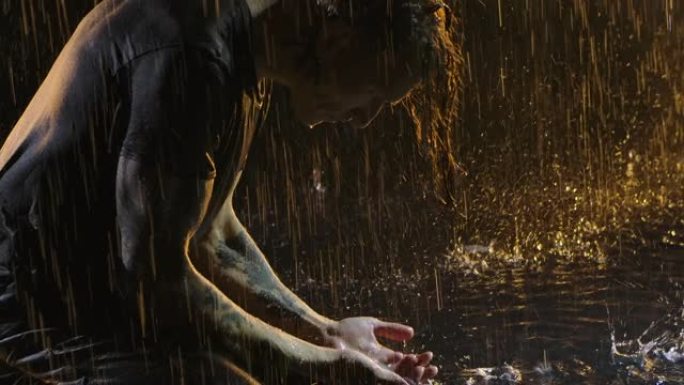 人手里拿着指南针，专心地盯着它。一个湿男人在倾盆大雨中跪在水面上。在黄色灯光照亮的黑暗工作室拍摄。特