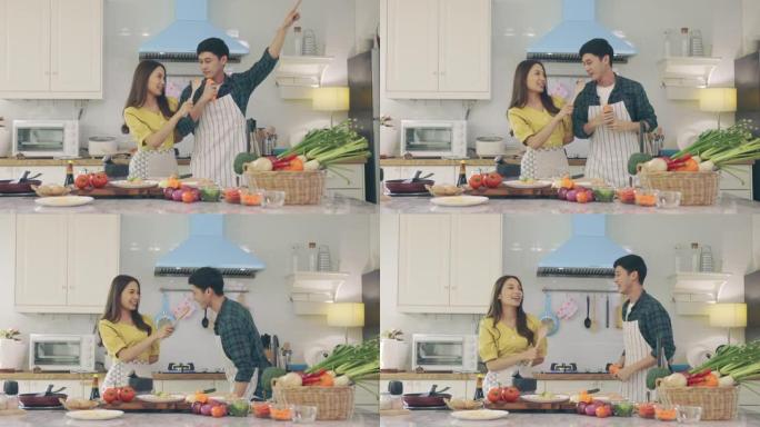 年轻的亚洲快乐活跃的家庭夫妇一起跳舞笑着在家准备食物，无忧无虑快乐的夫妻享受烹饪健康浪漫晚餐的乐趣在