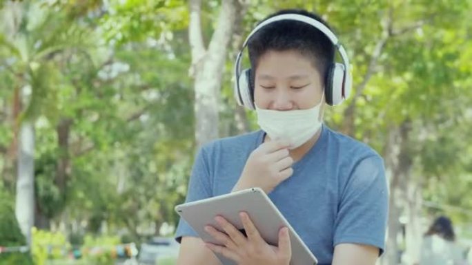 快乐的亚洲男生戴着口罩，在校园户外用数字平板电脑和耳机放松，社交疏远的生活方式理念。