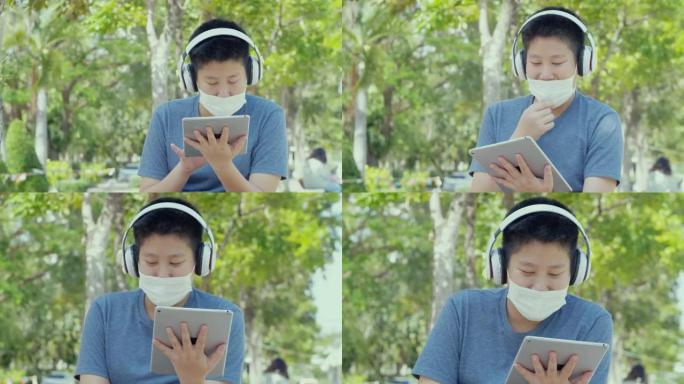 快乐的亚洲男生戴着口罩，在校园户外用数字平板电脑和耳机放松，社交疏远的生活方式理念。