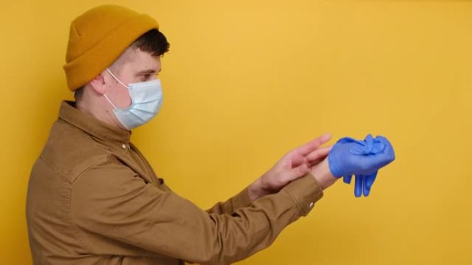 戴着医用口罩的年轻人的侧视图取下医用手套，扔进垃圾桶，穿着衬衫和无檐小便帽，隔离在黄色工作室背景下，