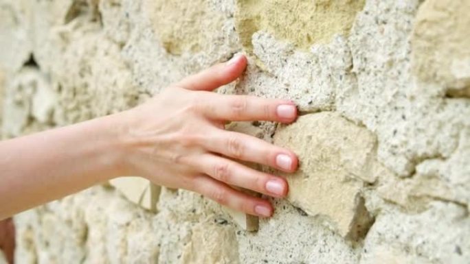女人在慢动作中滑手靠在旧石墙上。女性手触摸岩石粗糙表面