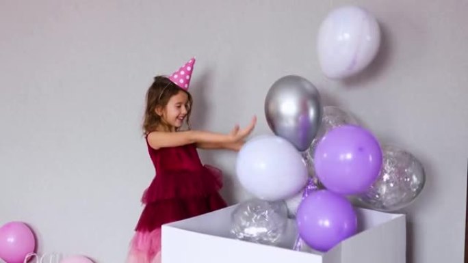 穿着粉色连衣裙和帽子的快乐小女孩在家里生日派对上打开带气球的大礼物礼品盒
