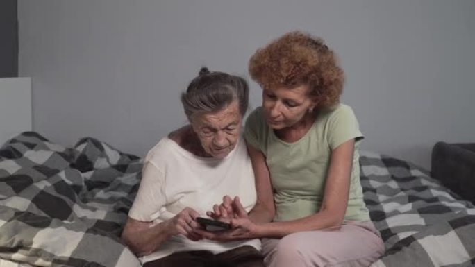 年迈的女儿教她的老母亲使用手机。成熟的护士帮助老年女性如何在疗养院使用智能手机。照顾者和老年人在床上